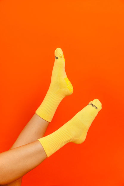 Un-banana organic cotton grip socks in Apricot Sorbet, worn with legs raised against a bright orange backdrop, sole grips visible.