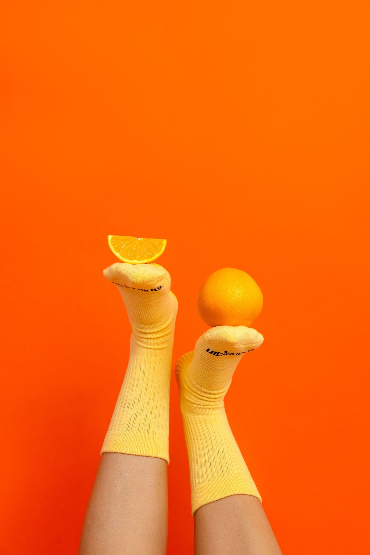 Un-banana organic cotton grip socks in Apricot Sorbet, legs raised against an orange backdrop, an orange balanced on one foot and a slice on the other.