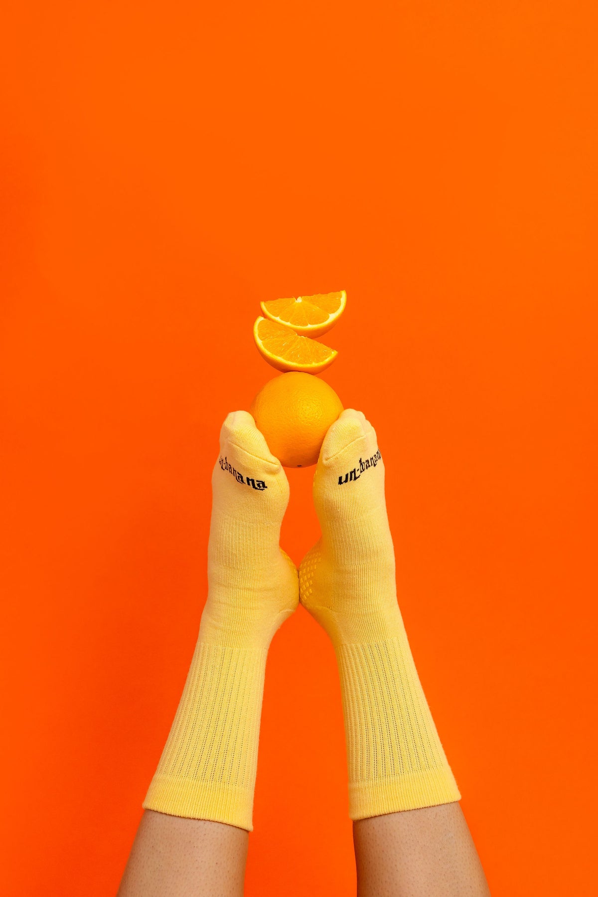 Un-banana organic cotton grip socks in Apricot Sorbet, feet pressed together with orange slices stacked on the toes against an orange backdrop.
