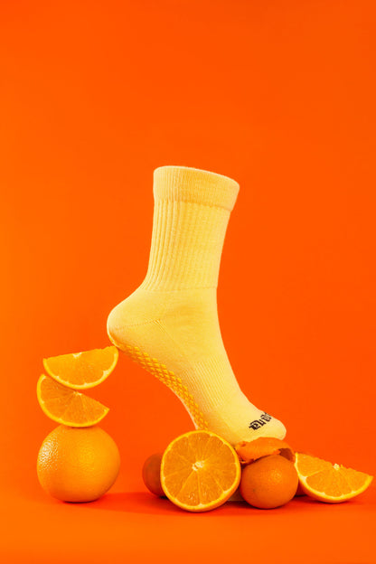 Un-banana Apricot Sorbet organic cotton grip sock posed on a pile of whole and sliced oranges, sole grips visible, on an orange backdrop.