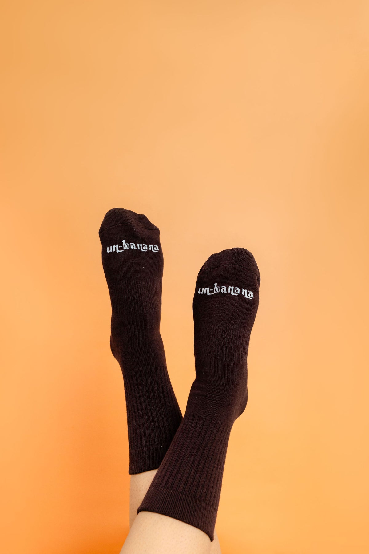 Un-banana organic cotton grip socks in Chocolate & Dates, legs raised and crossed against a warm apricot backdrop.