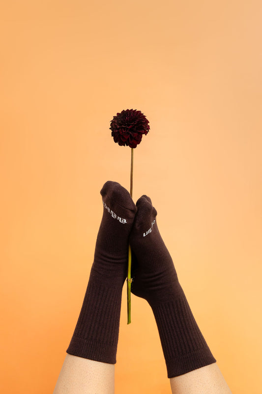 Un-banana organic cotton grip socks in Chocolate & Dates, feet raised holding a deep burgundy dahlia against a peach backdrop.
