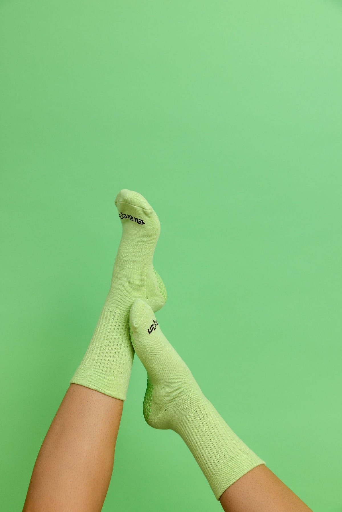 Un-banana organic cotton grip socks in Pistachio, legs raised and crossed against a mint green backdrop.