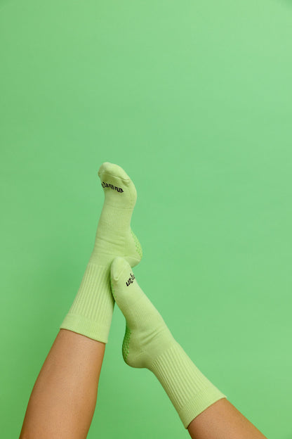 Un-banana organic cotton grip socks in Pistachio, legs raised and crossed against a mint green backdrop.