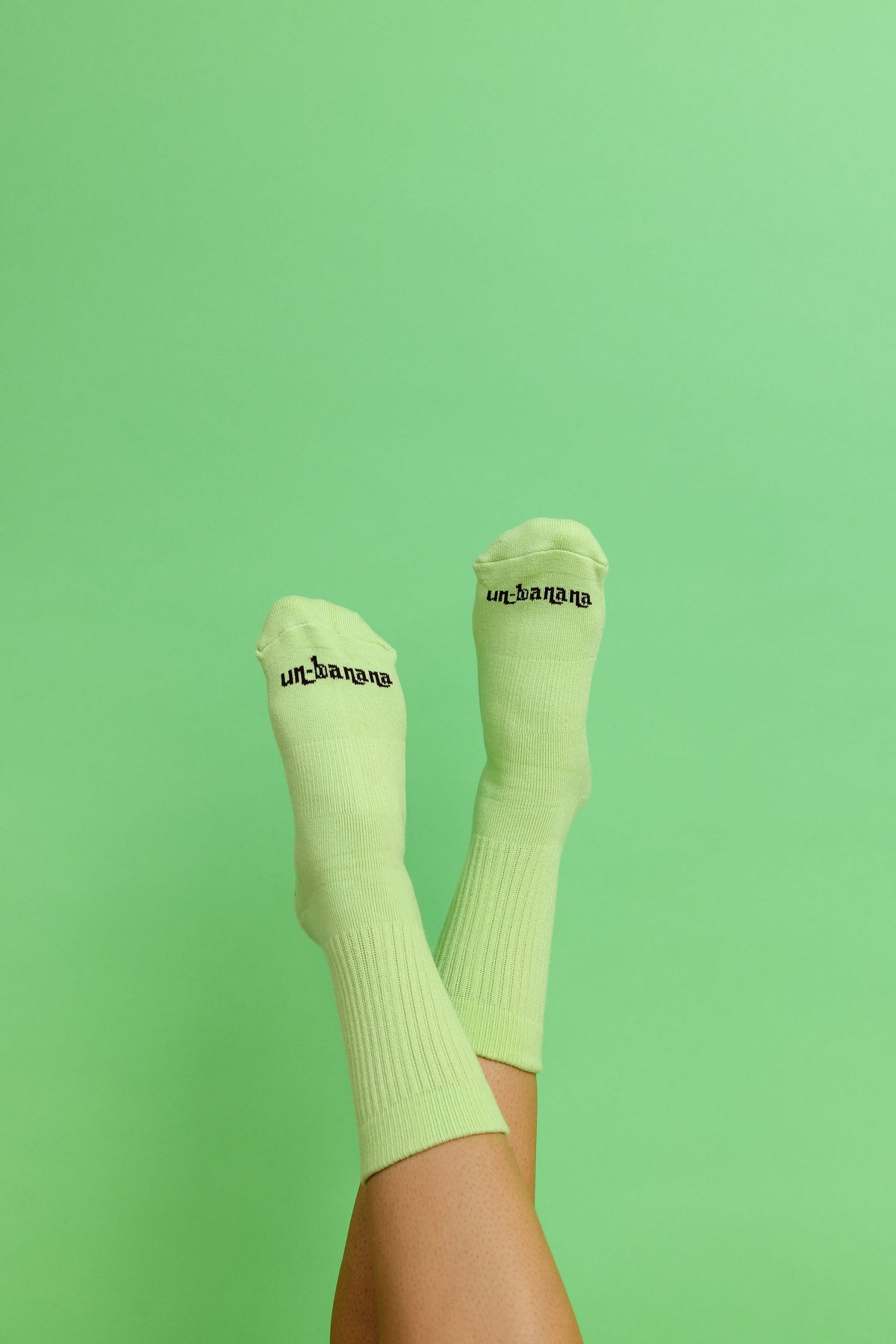 Un-banana organic cotton grip socks in Pistachio, legs raised side by side against a mint green backdrop, logo on the toes.