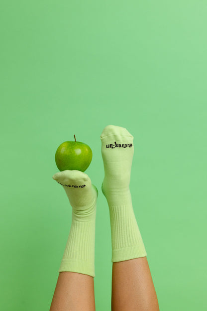 Un-banana organic cotton grip socks in Pistachio, feet raised with a green apple balanced on one foot against a mint green backdrop.
