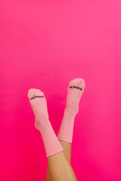 Un-banana organic cotton grip socks in Strawberry Milk, legs raised against a hot pink backdrop, logo on the toes.