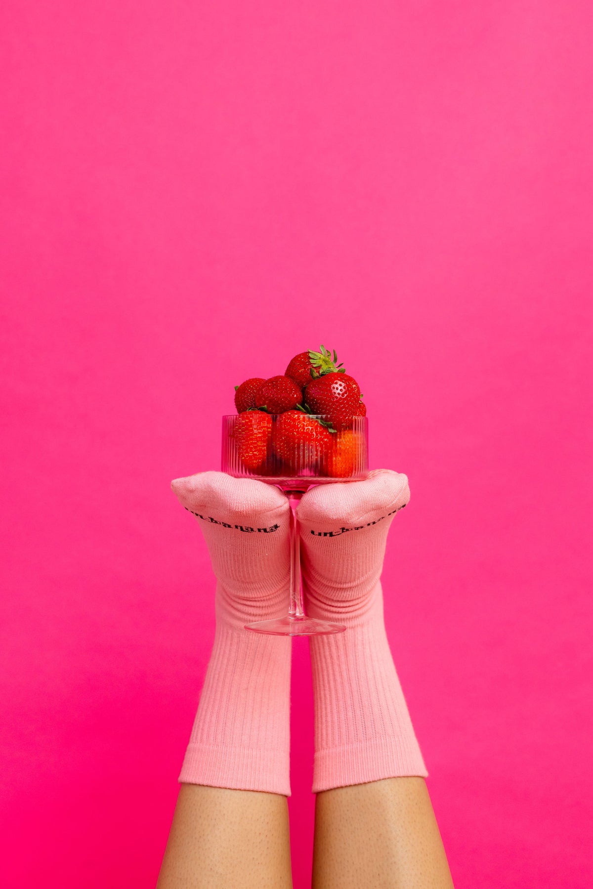 Un-banana organic cotton grip socks in Strawberry Milk, feet raised holding a clear container of strawberries against a hot pink backdrop.