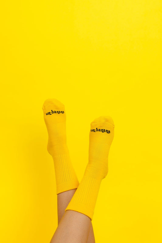 Un-banana organic cotton grip socks in The Peel, legs raised against a yellow backdrop, logo on the toes.