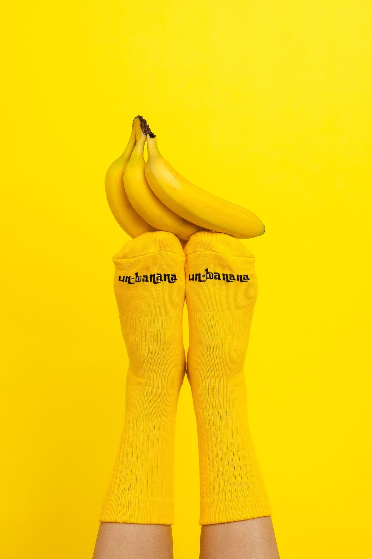 Un-banana organic cotton grip socks in The Peel, feet raised with a bunch of bananas balanced on the toes against a yellow backdrop.