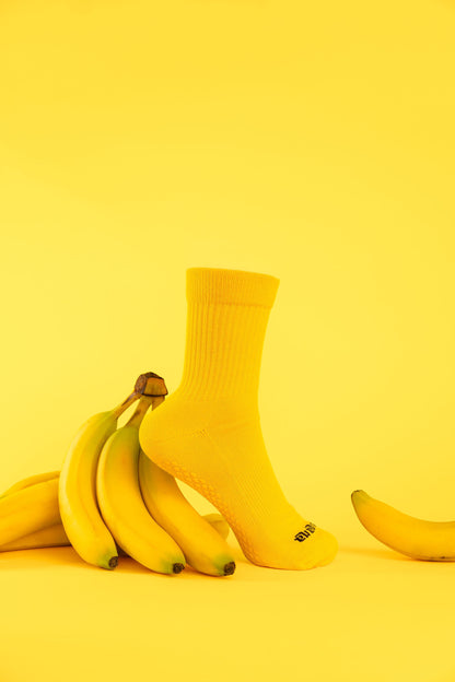 Un-banana organic cotton grip sock in The Peel standing upright beside a bunch of bananas on a yellow backdrop.
