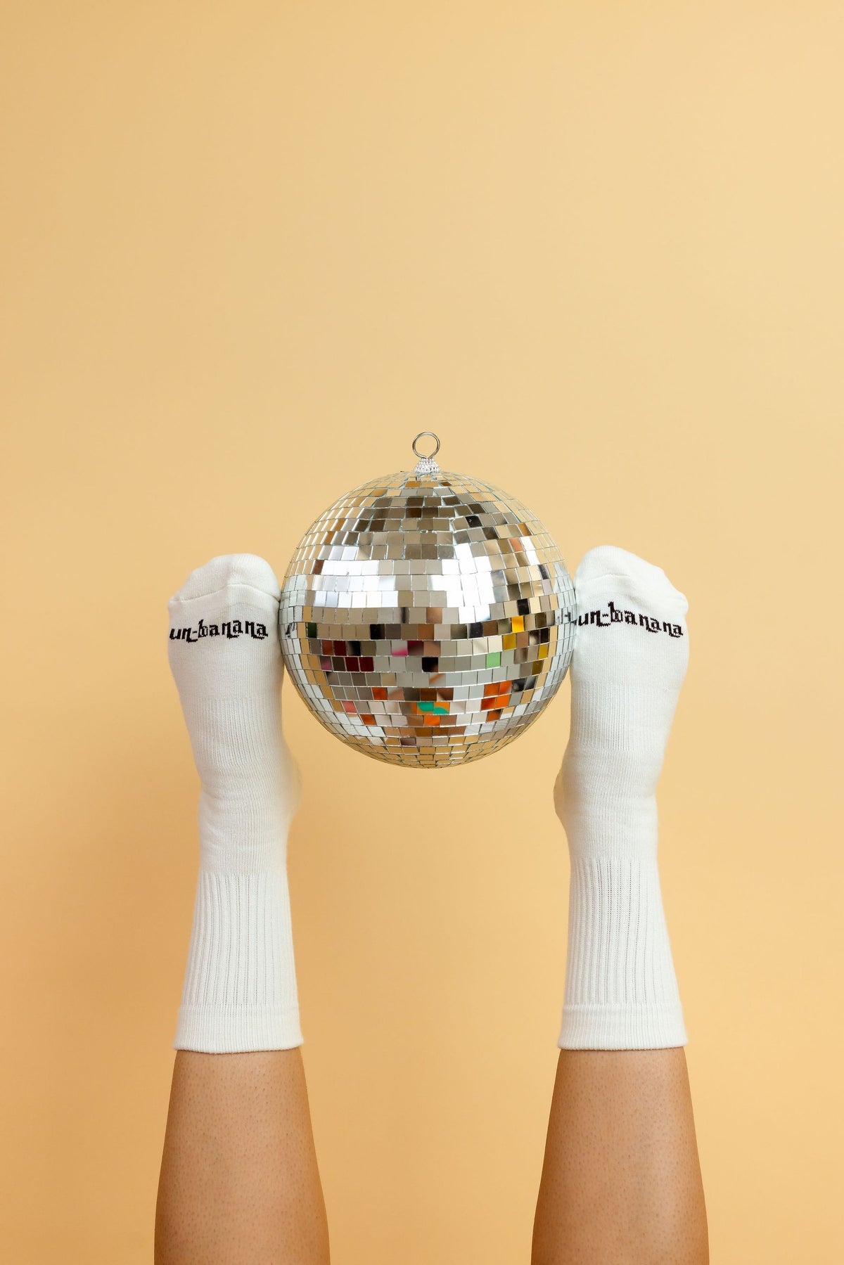 Un-banana organic cotton grip socks in Vanilla Bean, feet raised and holding a disco ball between them against a peach backdrop.