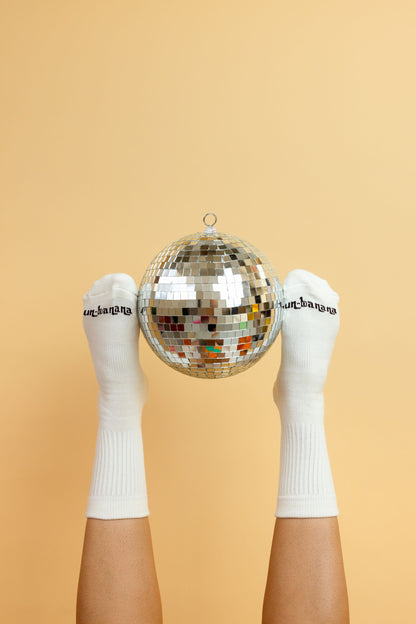 Un-banana organic cotton grip socks in Vanilla Bean, feet raised and holding a disco ball between them against a peach backdrop.