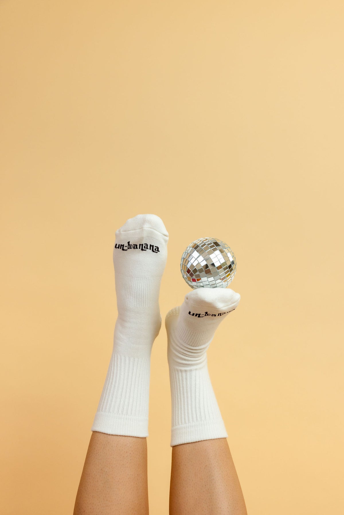 Un-banana organic cotton grip socks in Vanilla Bean, legs raised with a small disco ball balanced on one foot against a peach backdrop.