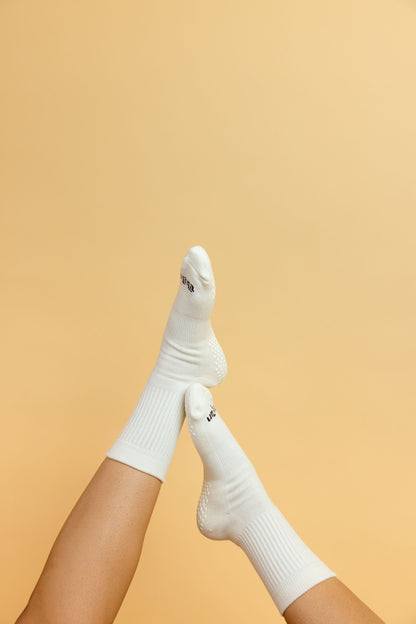 Un-banana organic cotton grip socks in Vanilla Bean, legs raised and crossed against a soft peach backdrop, sole grips visible.