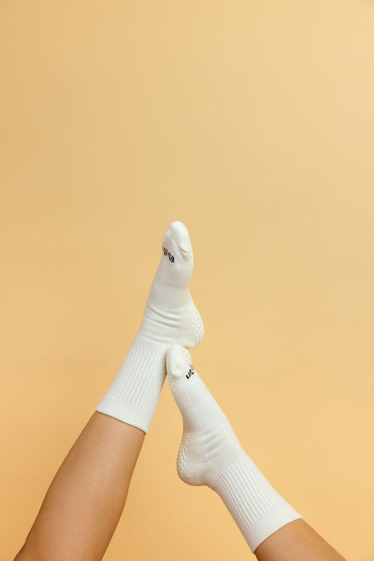 Un-banana organic cotton grip socks in Vanilla Bean, legs raised and crossed against a soft peach backdrop, sole grips visible.